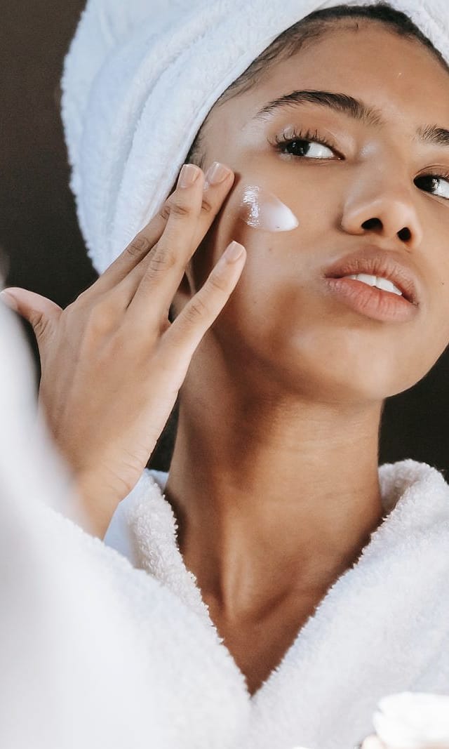 A woman with her hair wrapped in a towel dabs cream onto her cheek in soft daylight.