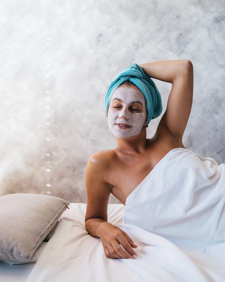 A woman in a clay face mask and a towel turban rests against white bedding.