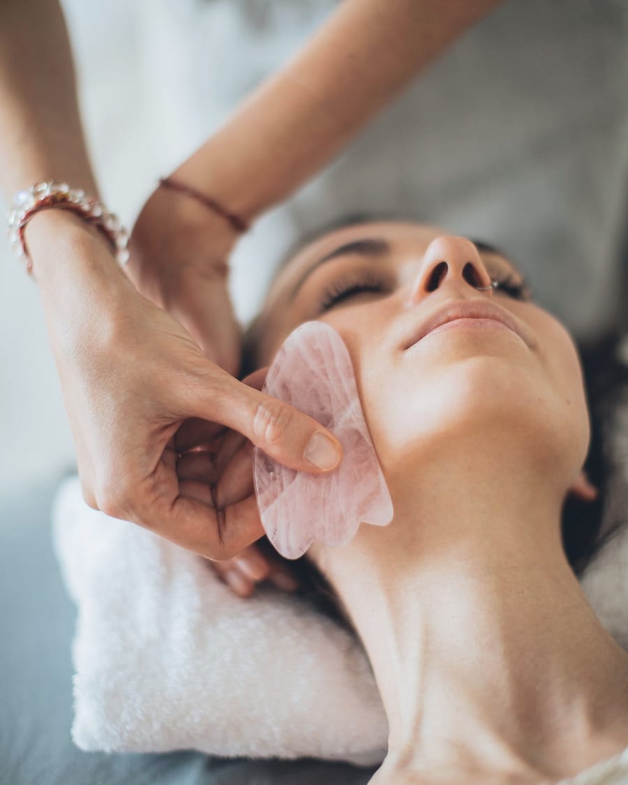 A practitioner's hands move a smooth stone gently along a client's jaw.