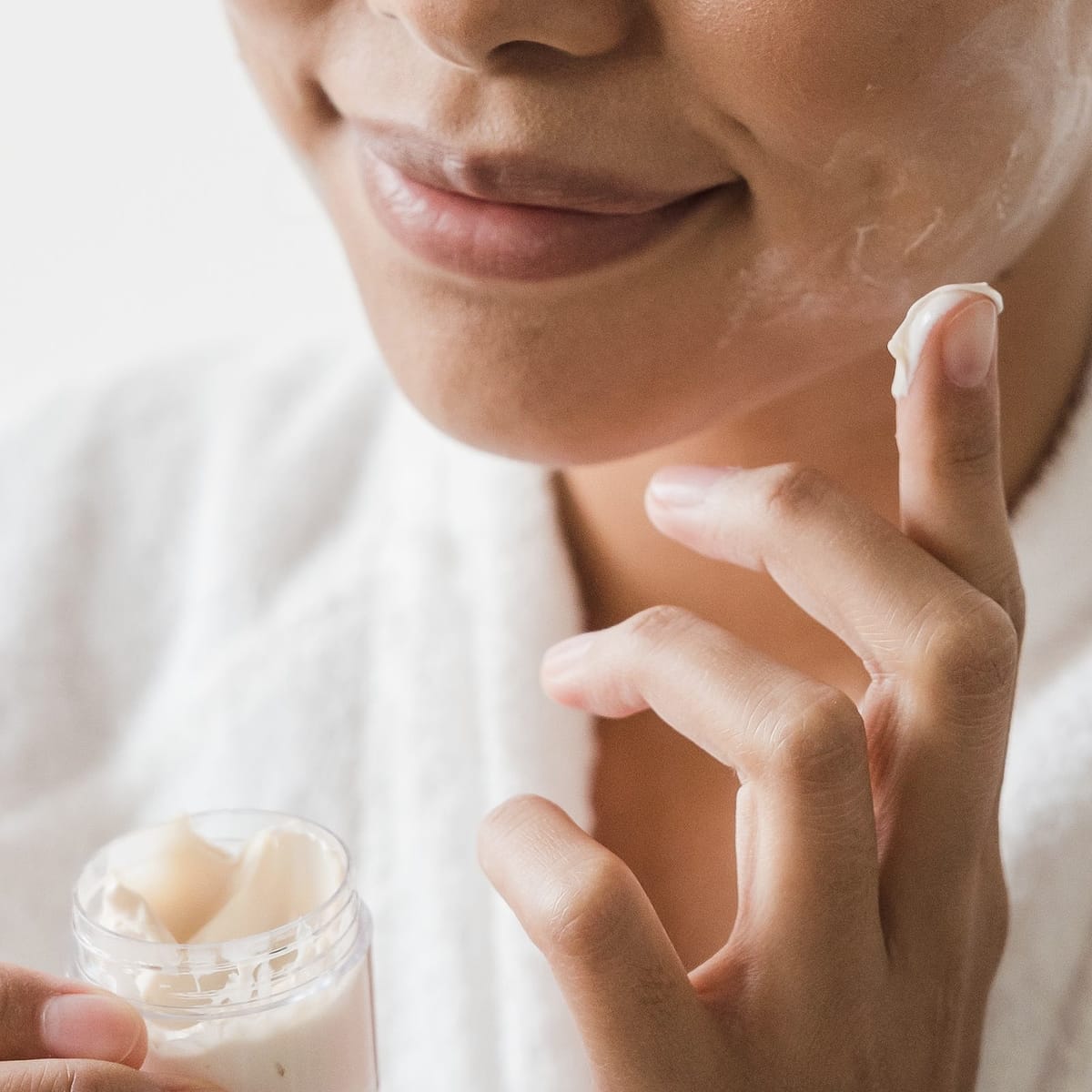 A woman in a white robe smooths a little cream onto her cheek, holding the open jar.